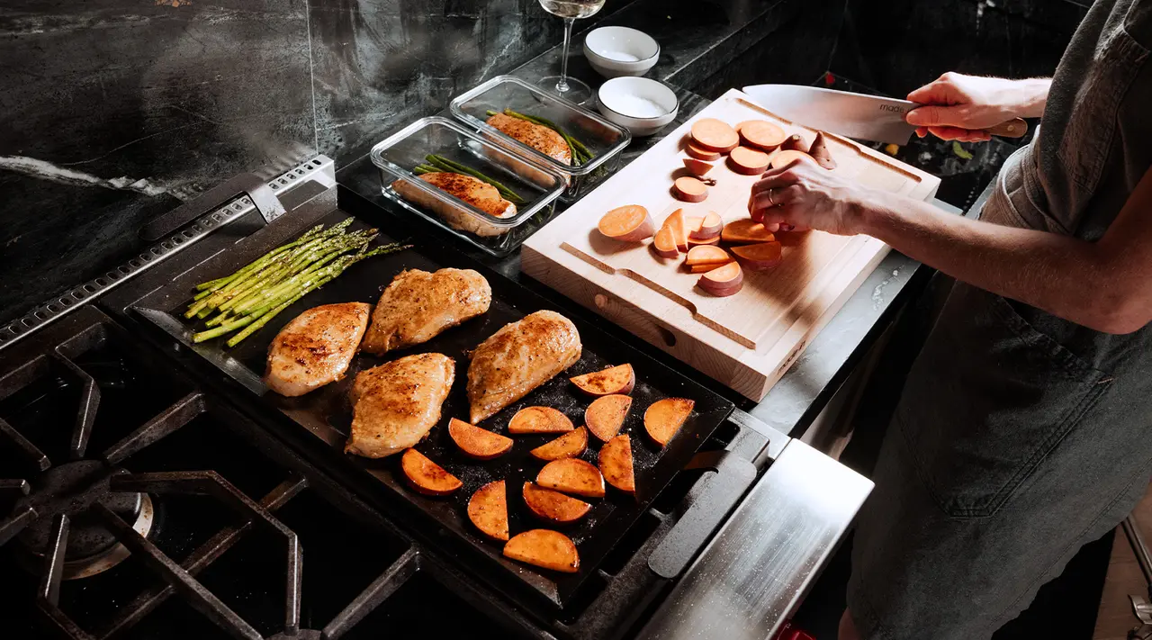 A person in an apron is cooking a meal with chicken breasts on a stove and cutting sweet potatoes on a cutting board in a kitchen.