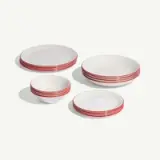 A set of white dishware with red rims, including plates and bowls, is arranged on a light surface.