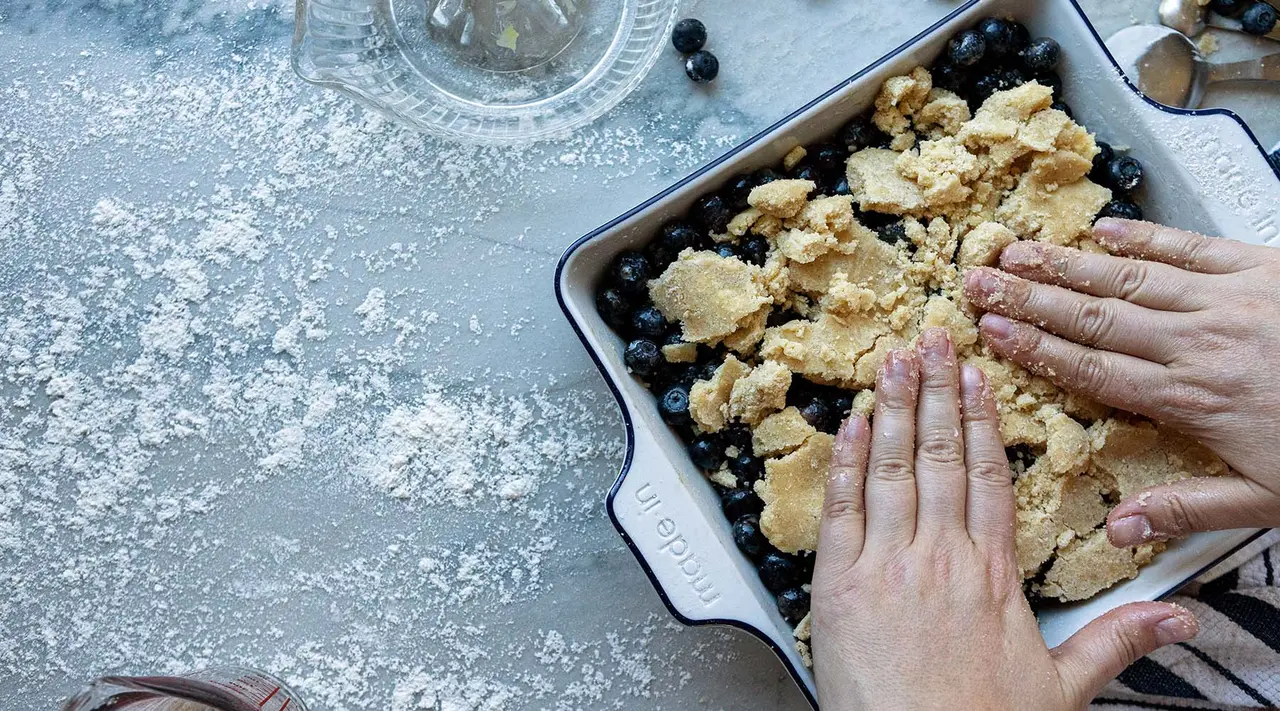 A person's hands are pressing crumbled dough into a baking dish with blueberries, on a flour-dusted surface indicating preparation of a dessert.