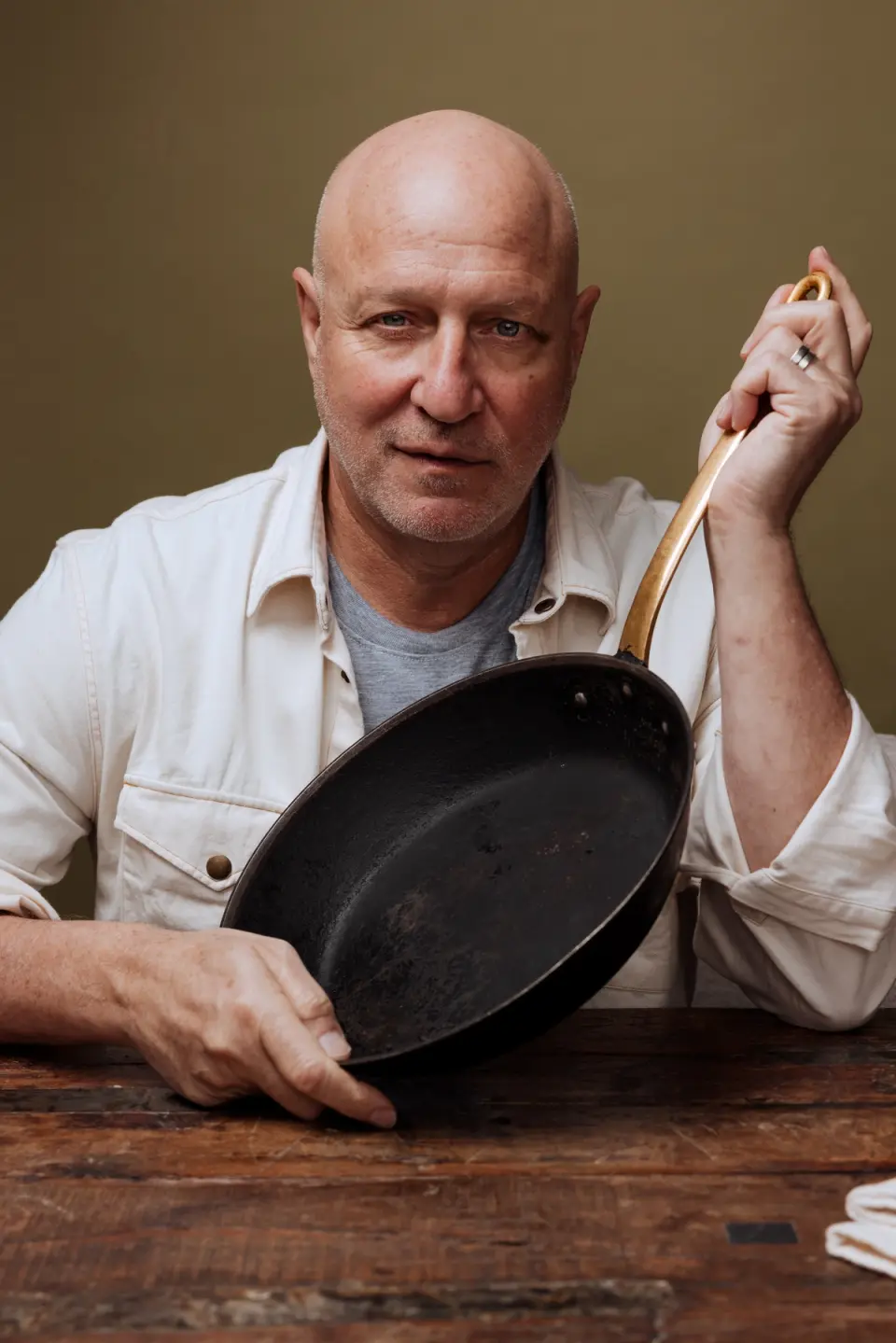 A bald man in a light-colored shirt confidently holds a frying pan while seated at a wooden table against a greenish background.