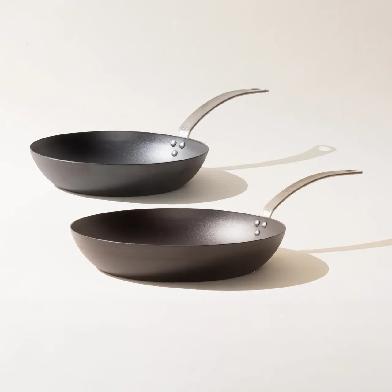 Two black non-stick frying pans with silver handles arranged diagonally on a light background.