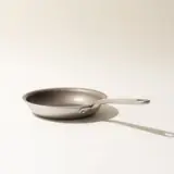 A stainless steel frying pan with a long handle is centered on a neutral background with a soft shadow indicating directional light.