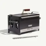 A compact, black stovetop grill with a stainless steel cooking surface and a wooden handle, accompanied by a removable tool for handling.