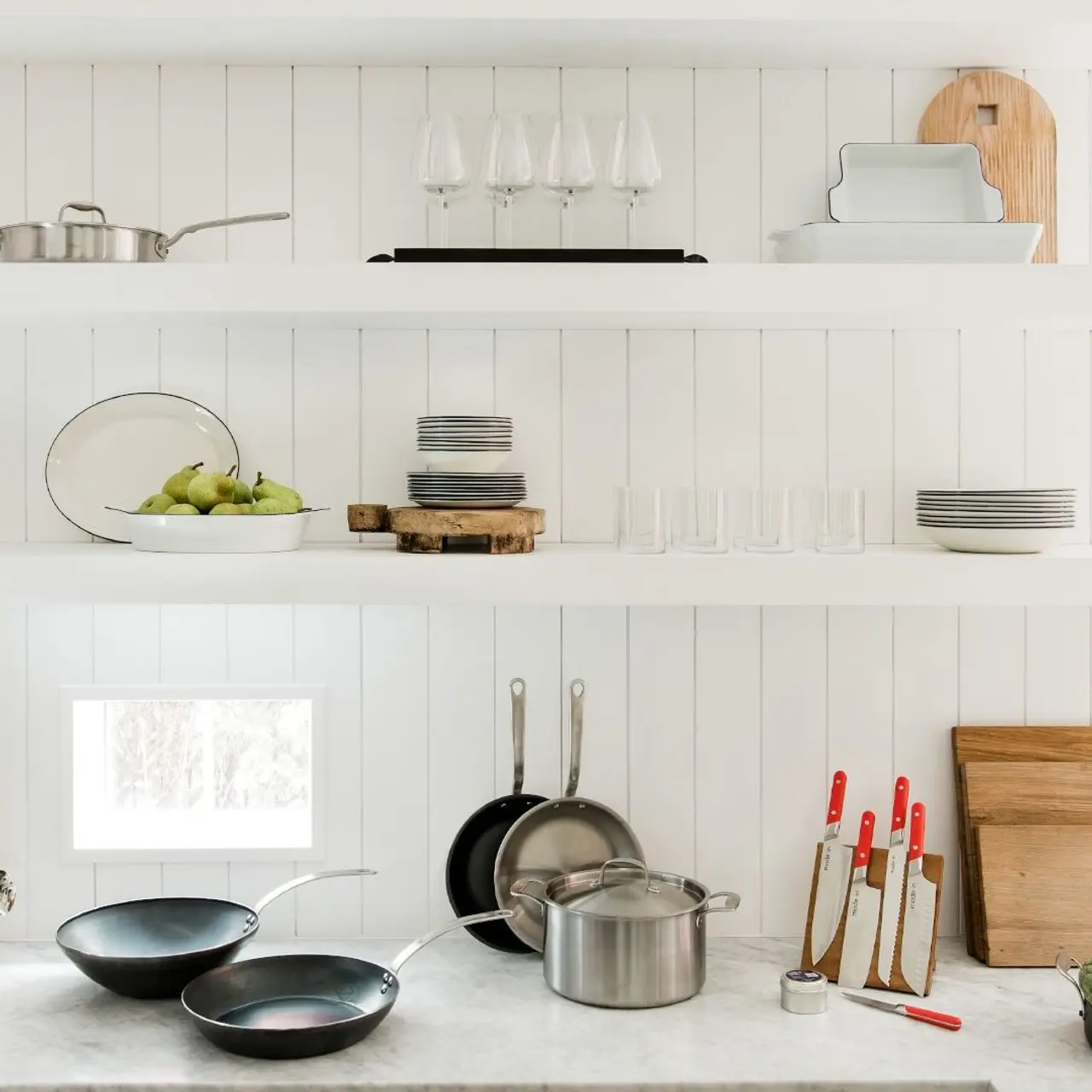 A minimalist kitchen shelf displays a variety of cookware, glassware, and decor against a bright white backdrop.