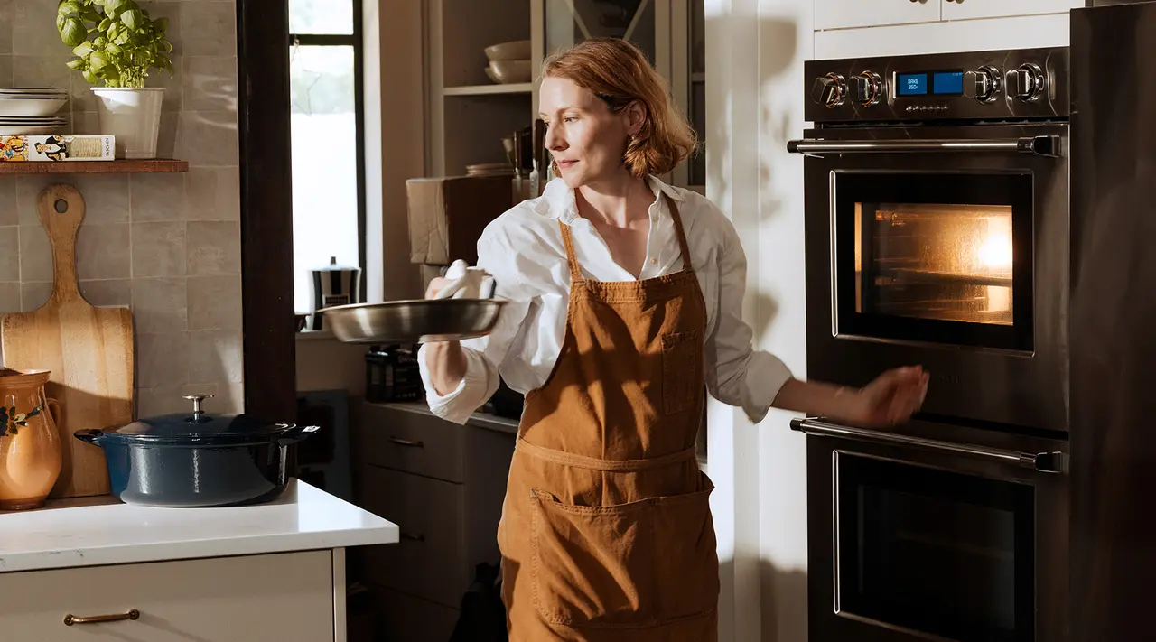 A person wearing an apron holds a pan with one hand while cooking in a well-equipped kitchen, expressing a cheerful demeanor.