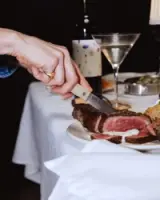 A hand holds a knife poised to cut a steak on a white tablecloth, with a glass of martini and a bottle of wine in the background.
