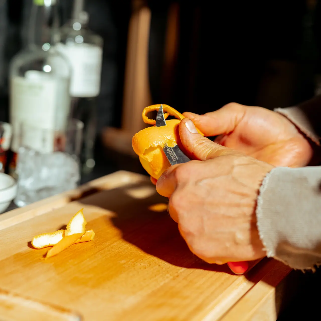 Hands peeling an orange with a knife on a wooden cutting board with peelings beside and bottles in the blurred background.