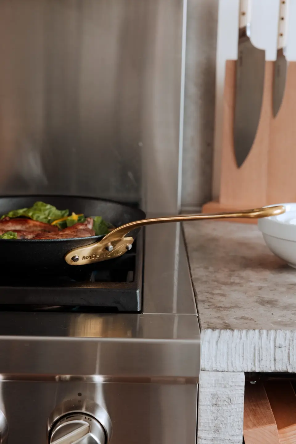 A steak with greens being cooked in a skillet on a stove, with a gold-tone pair of tongs resting on the edge.