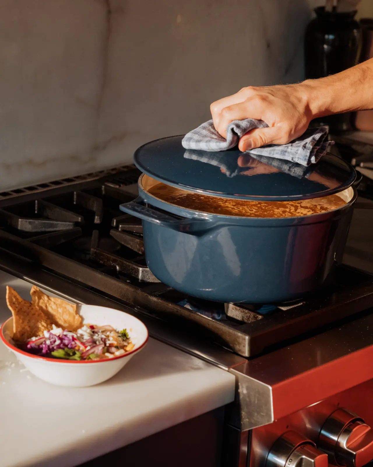 A person is lifting the lid of a large blue pot on a stove with a bowl of food garnished with onion and herbs and a tortilla chip in the foreground.