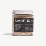 A clear glass jar labeled "MADE IN x" contains smoky onion salt, designed for enhancing flavors during summer grilling, with a net weight of 2.32 oz.
