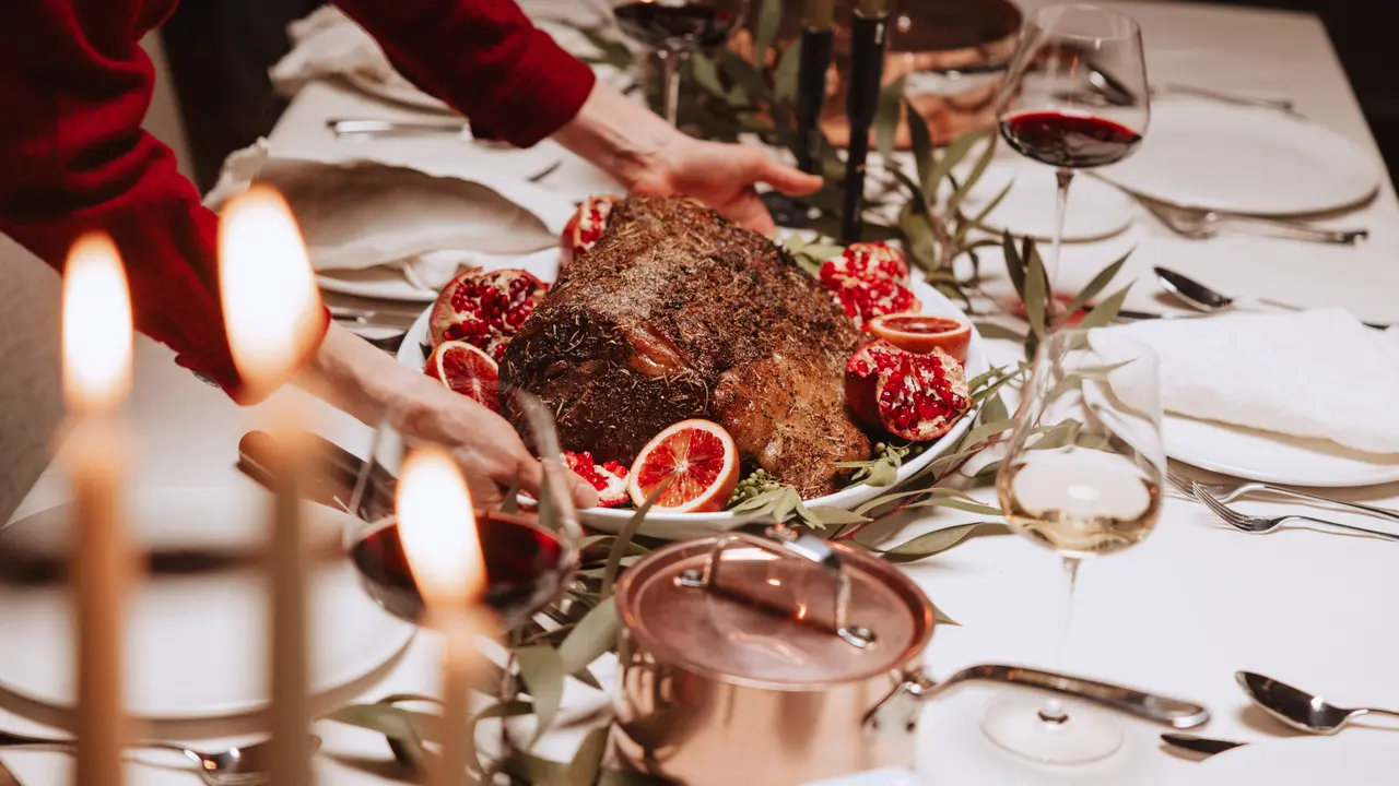 A beautifully set dining table features a person serving a roasted dish surrounded by pomegranates and elegant wine glasses.