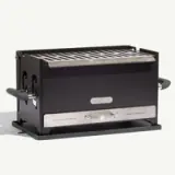 A compact, black portable grill with a stainless steel cooking grate and side handles, featuring a "Made-In" label.