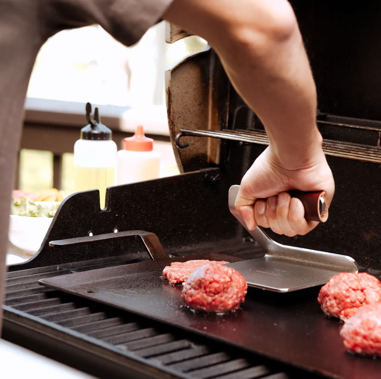 A person's arm is visible as they press down on hamburger patties with a spatula on an outdoor grill.