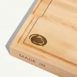 A wooden cutting board features a rounded logo in the corner and the words "MADE IN" engraved on the side.
