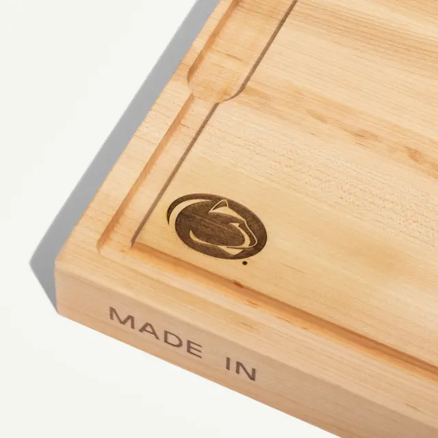 A wooden cutting board features a rounded logo in the corner and the words "MADE IN" engraved on the side.