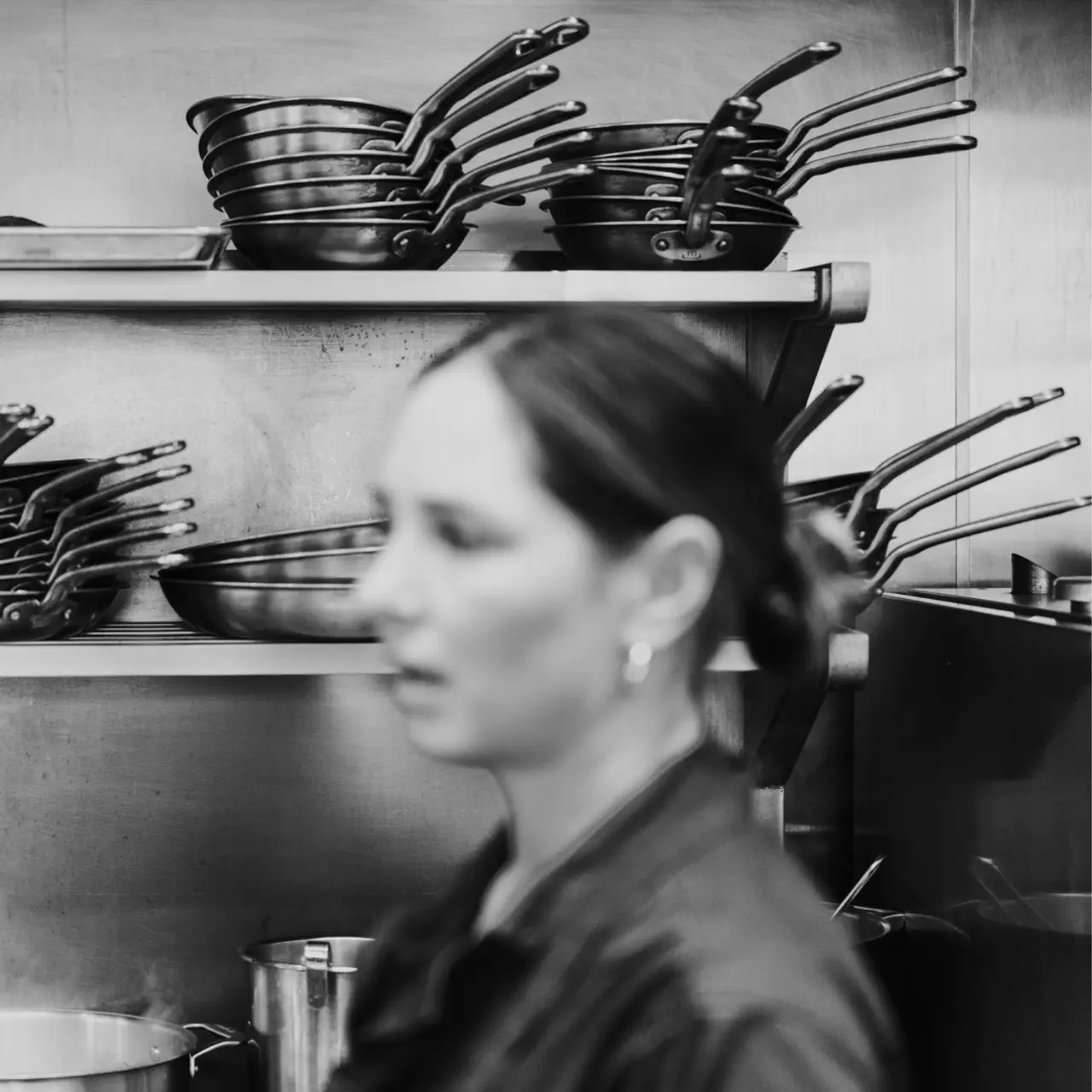 A woman stands in a kitchen with shelves of pans blurred in the background, emphasizing her motion or the hectic environment.