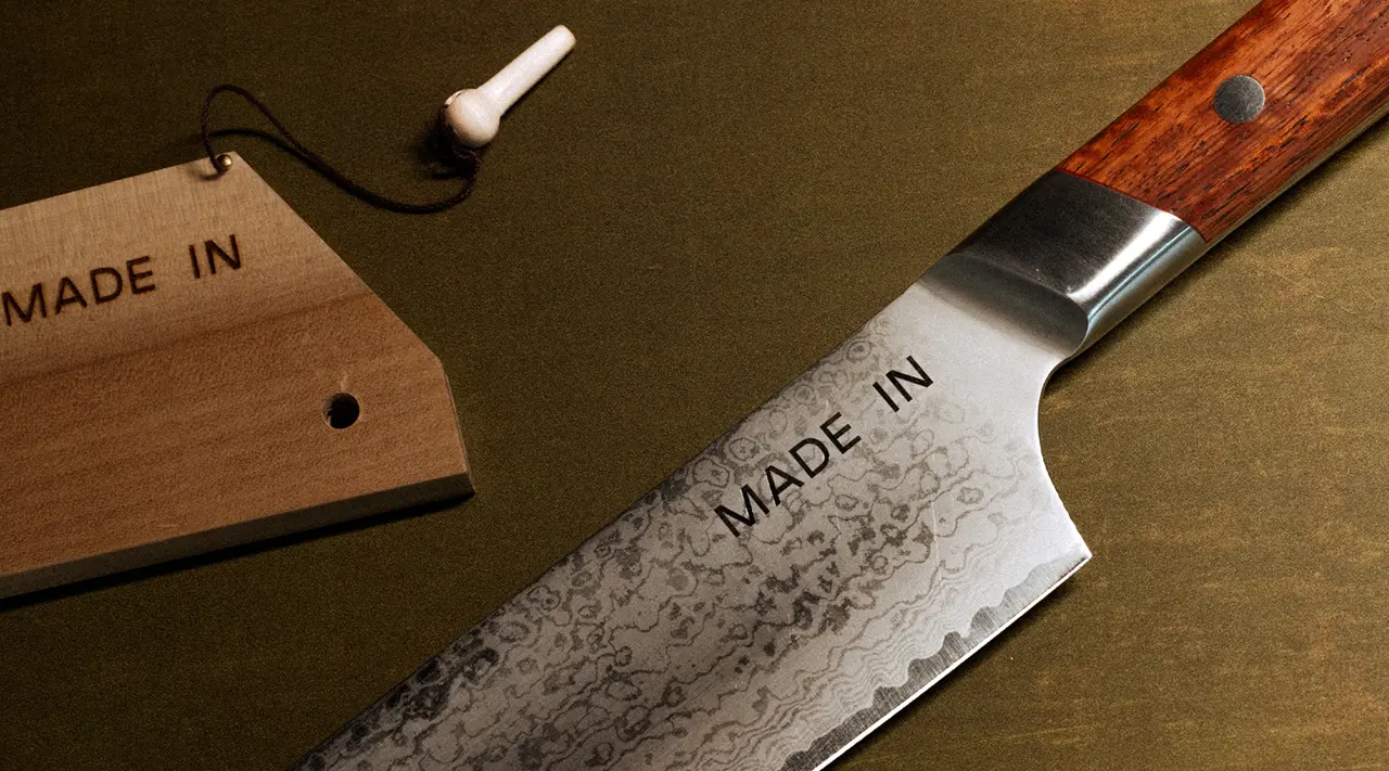 A chef's knife with "MADE IN" etched on the blade lies next to a wooden tag with the same inscription, implying a focus on craftsmanship origin.
