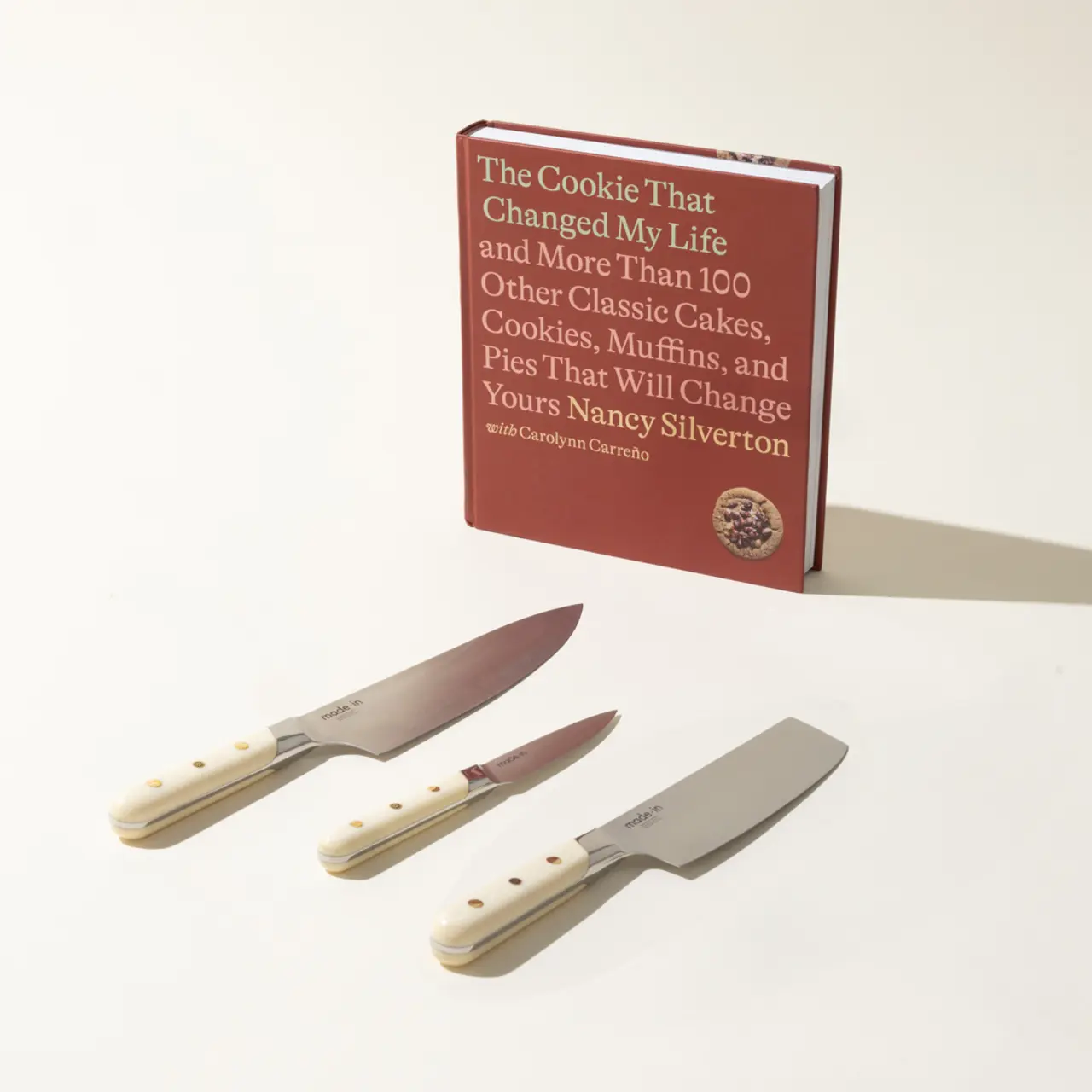 A cookbook titled "The Cookie That Changed My Life" is displayed upright behind three kitchen knives with cream-colored handles on a light surface.