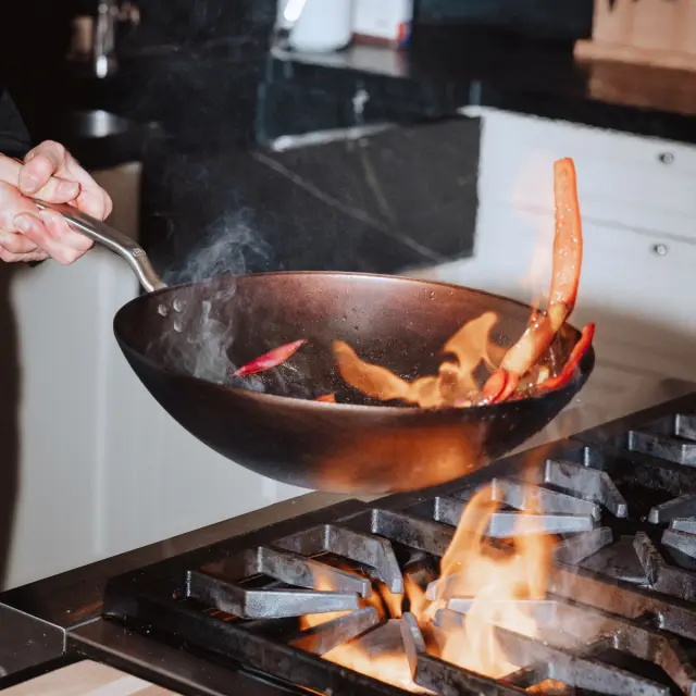 A person is skillfully tossing vegetables in a sizzling wok over a gas stove, with flames and smoke creating a dynamic cooking scene.