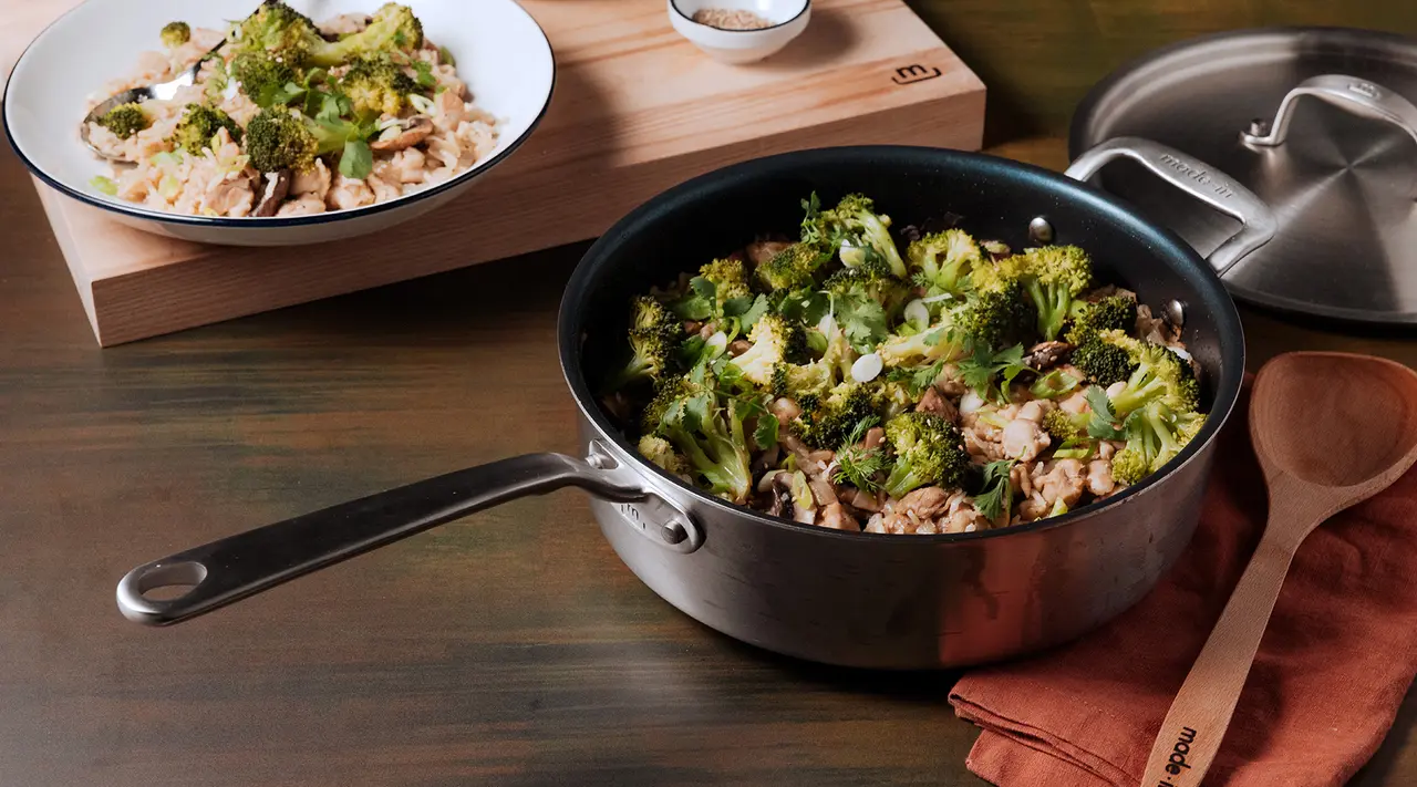 A skillet with broccoli and chunks of meat is placed on a wooden counter alongside a plate of similar food content, a pot lid, and a wooden spoon on a napkin.