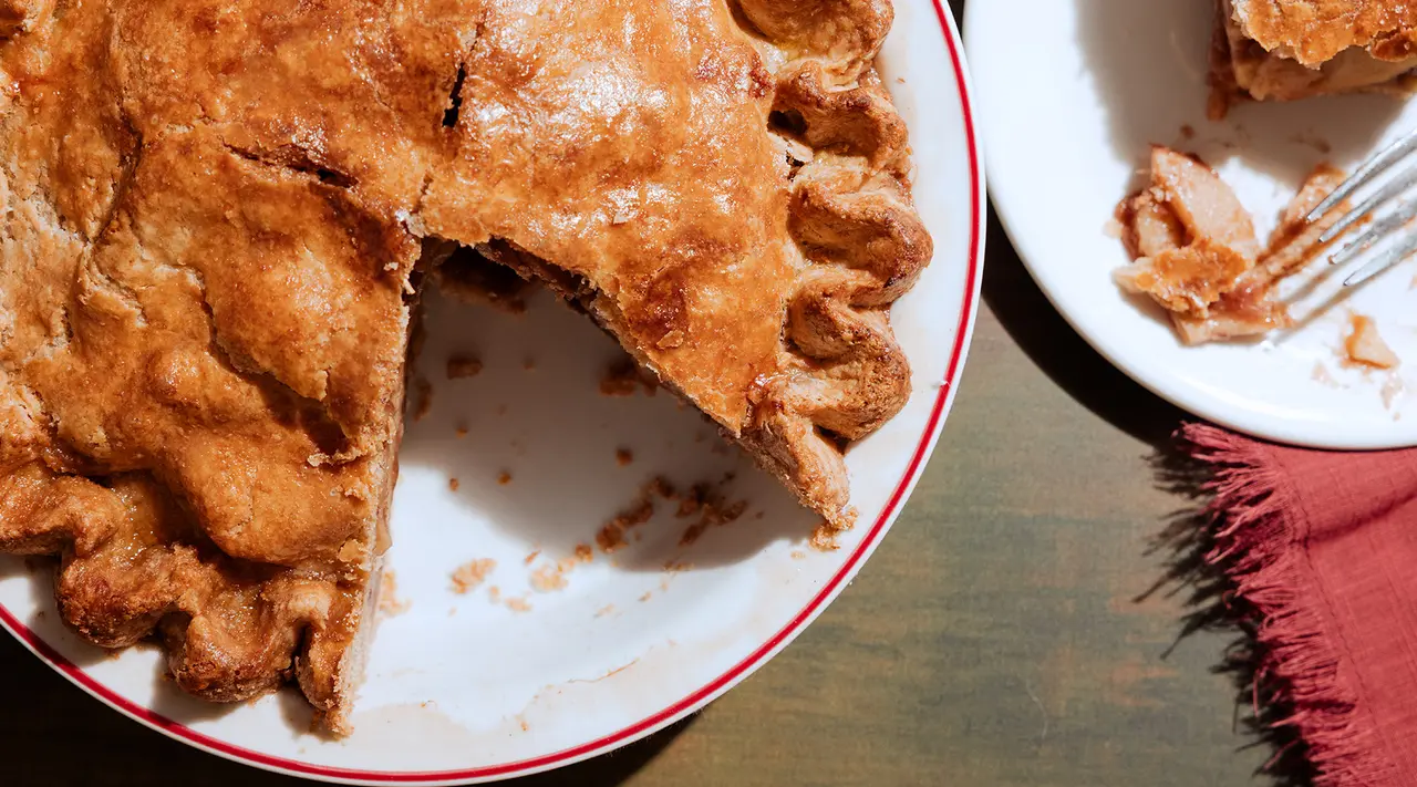 A partially eaten pie with a flaky golden crust is on a plate alongside a smaller plate with a few crumbs.