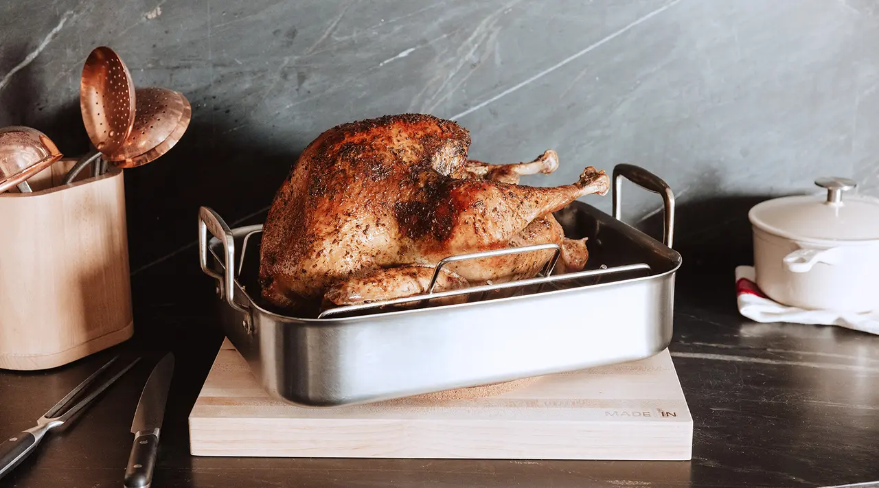 A roasted turkey sits in a stainless steel pan on a wooden cutting board, surrounded by cooking utensils.