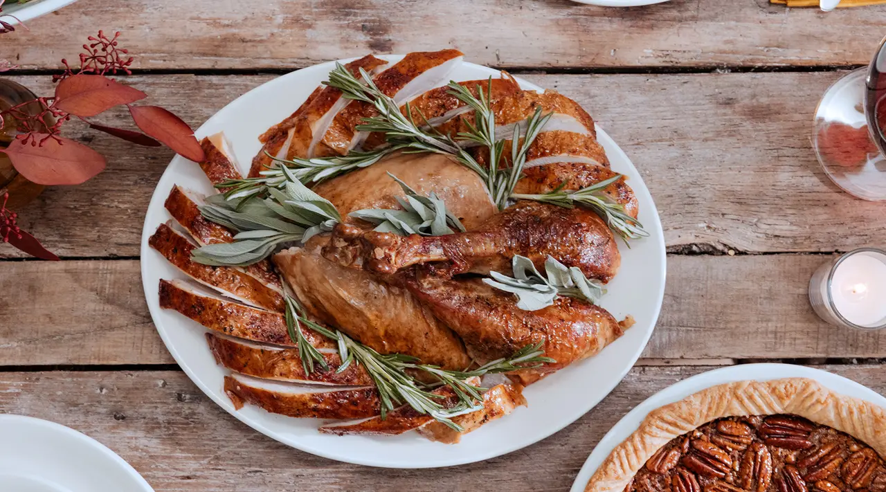 Roasted turkey garnished with herbs presented on a platter on a wooden table accompanied by side dishes and a pie, creating a festive meal setting.