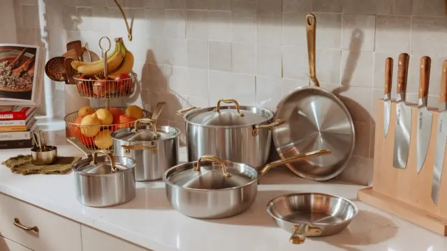 A collection of shiny stainless steel pots and pans, along with fresh fruit in a wire basket and a set of kitchen knives, are neatly arranged on a kitchen countertop.