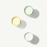 Three colorful candles in yellow, green, and blue containers casting shadows on a white surface.