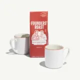 A bag of Founders' Roast whole bean coffee is displayed between two white mugs with red rims.