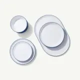 An arrangement of white ceramic dinnerware including bowls, plates, and platters with blue rims is laid out on a plain background.