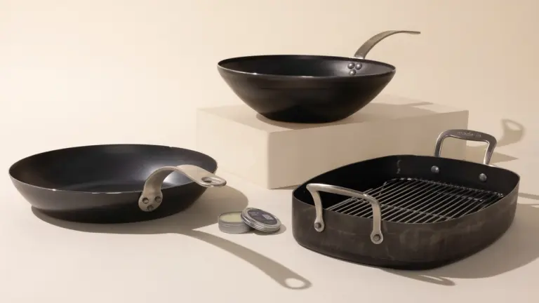Three different types of black frying pans displayed on a light background with one elevated on a beige block, featuring a variety of handles and one with a grill insert.