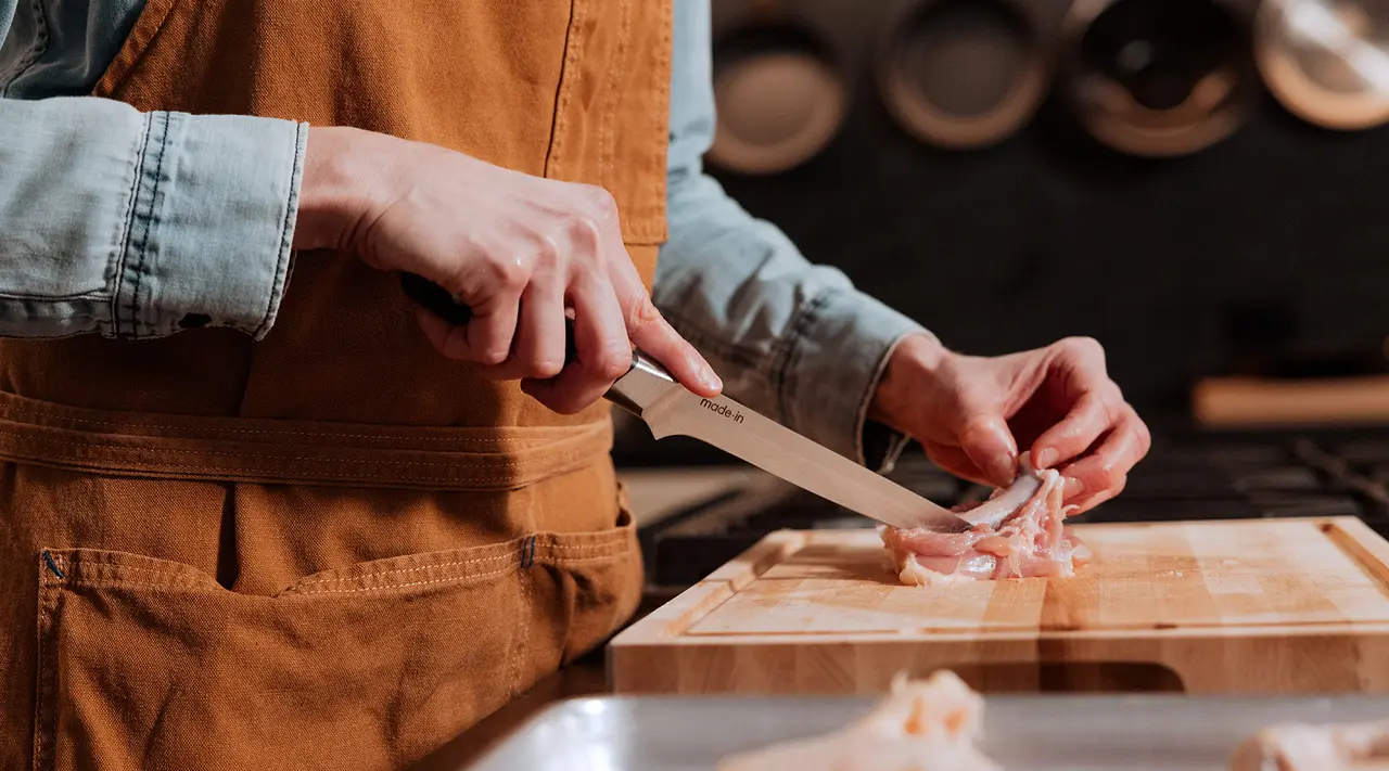 A person wearing an apron is cutting raw chicken on a wooden chopping board in a kitchen setting.