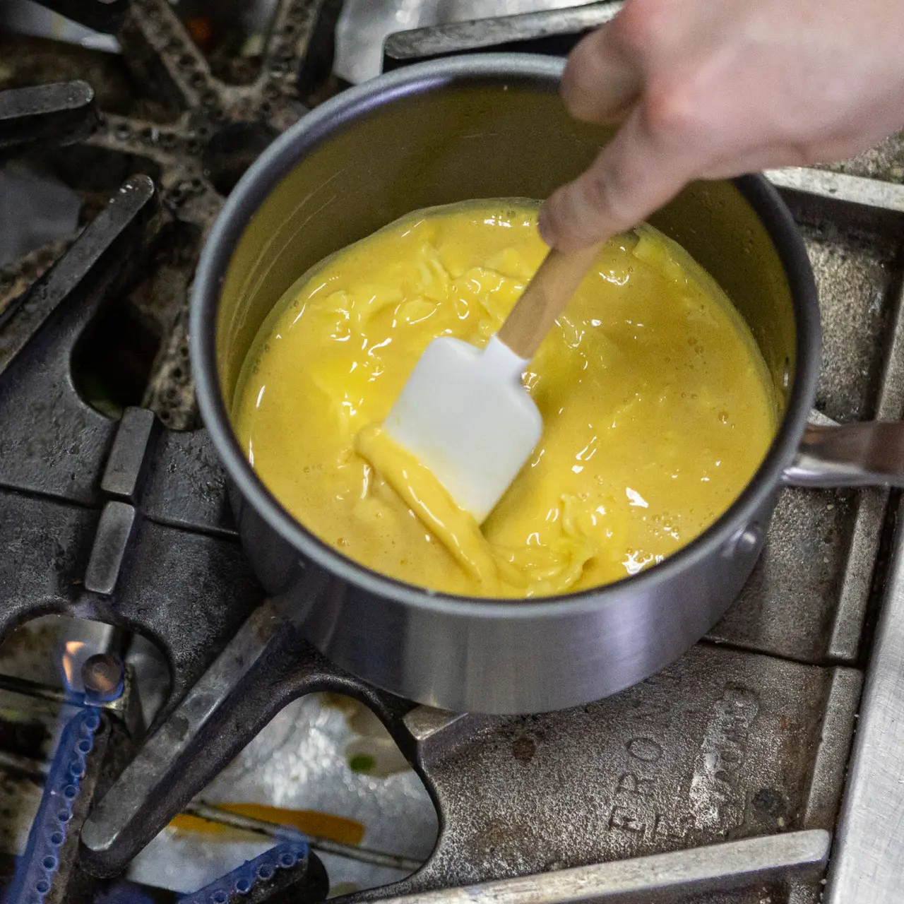 A person is stirring a thick yellow mixture in a saucepan on a stove with a white spatula.