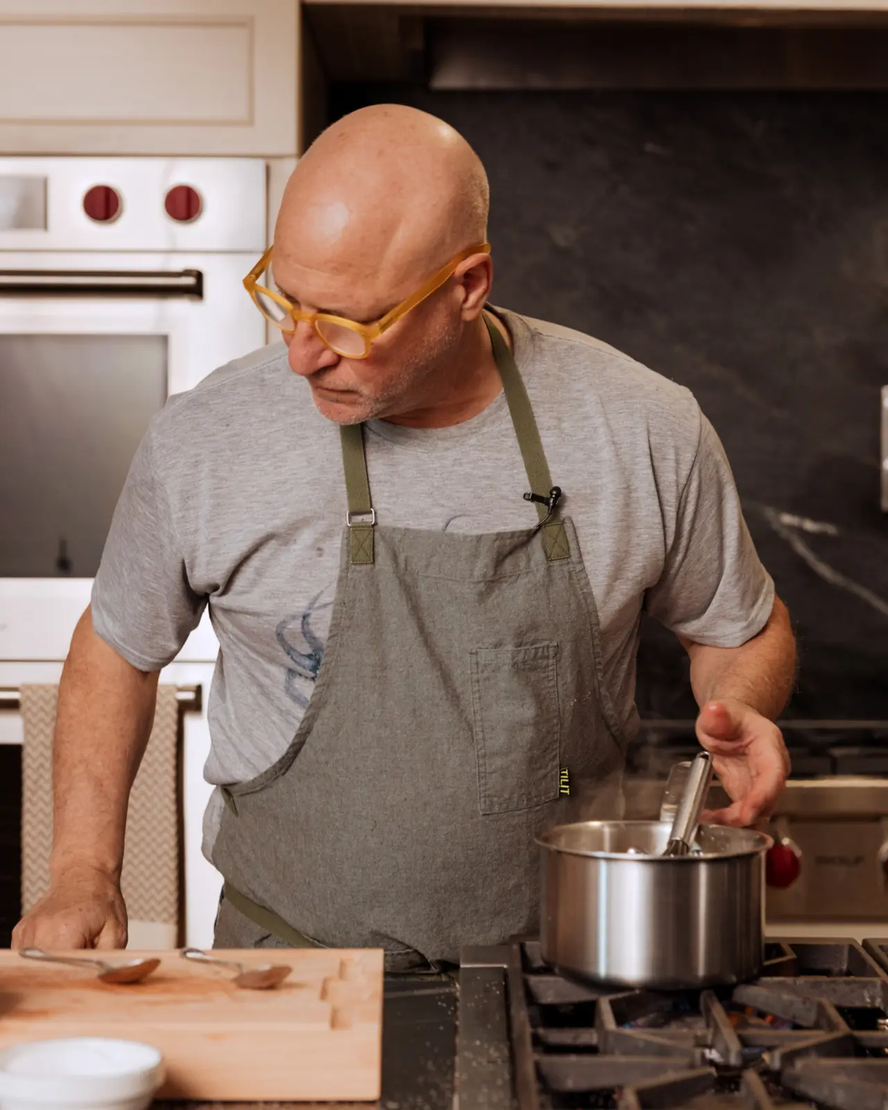 A bald man wearing eyeglasses and an apron stirs a pot on the stove while concentrating on his cooking.