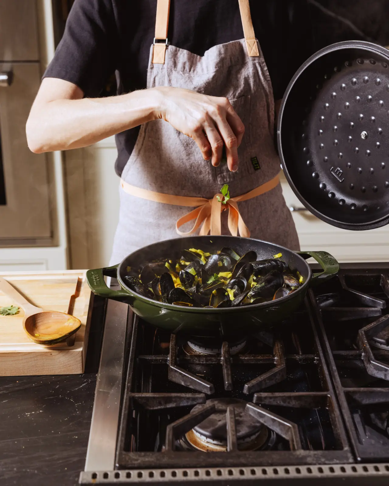 A person wearing an apron is preparing mussels in a green pot on a stovetop, adding seasoning from a lid.