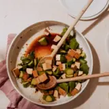 A bowl of diced tofu and sliced cucumbers with a pair of chopsticks rests on a table with a pink cloth and an empty small plate nearby.