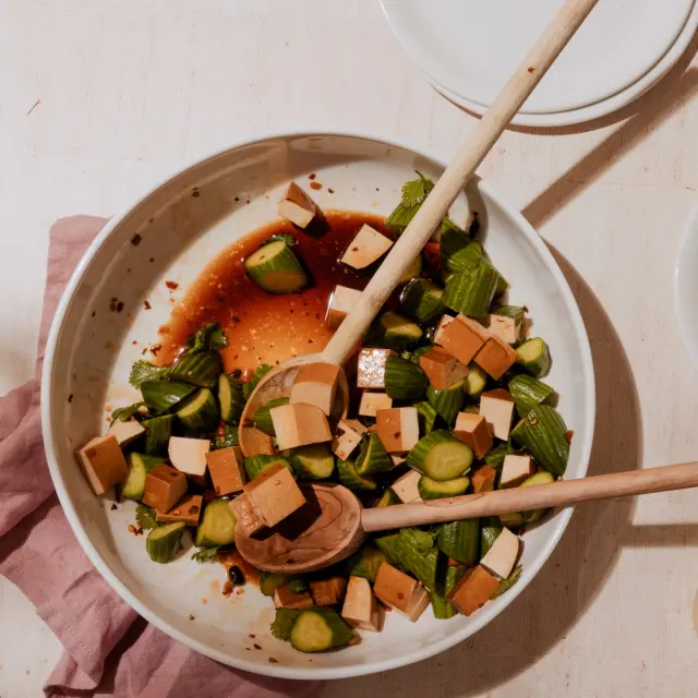 A bowl of diced tofu and sliced cucumbers with a pair of chopsticks rests on a table with a pink cloth and an empty small plate nearby.