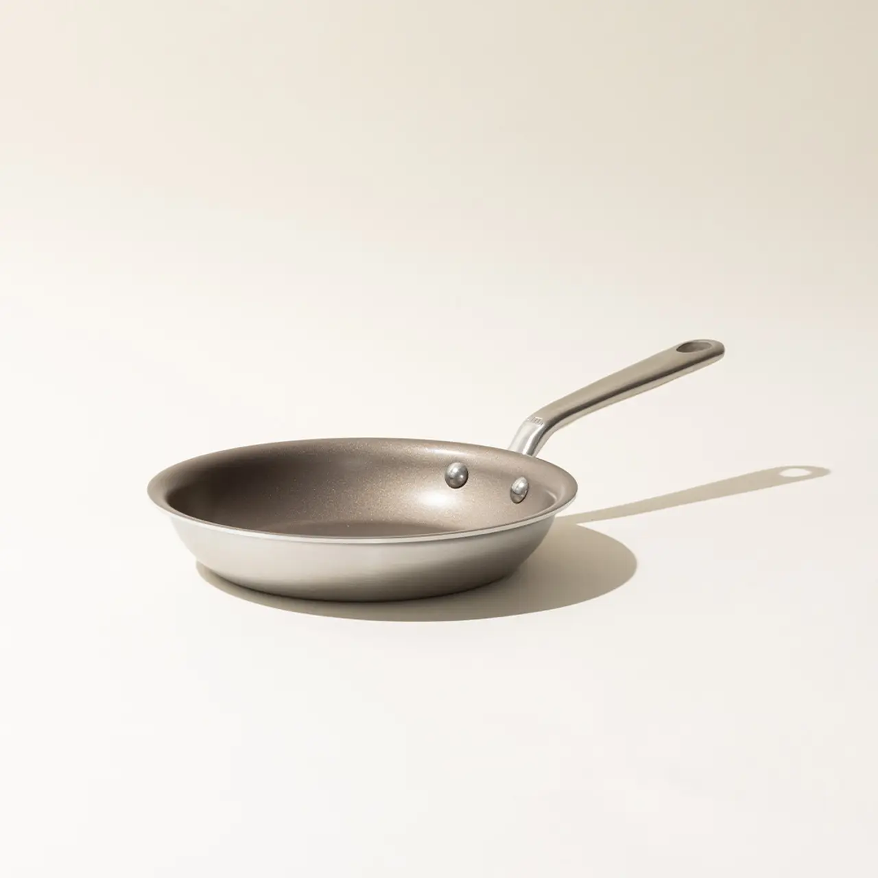 A single stainless steel frying pan on a neutral background with a shadow indicating studio lighting.