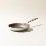 A single stainless steel frying pan on a neutral background with a shadow indicating studio lighting.