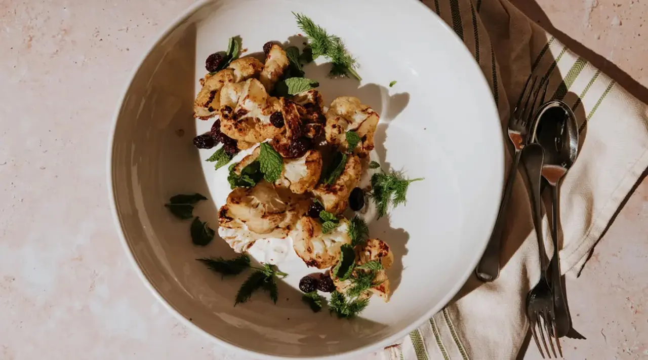 A plate of roasted cauliflower with herbs and raisins, accompanied by silverware on a textured table.