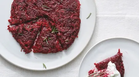 A round beetroot dish, cut into wedges, is garnished with sprigs of herbs and accompanied by a serving on a separate plate topped with a dollop of cream.