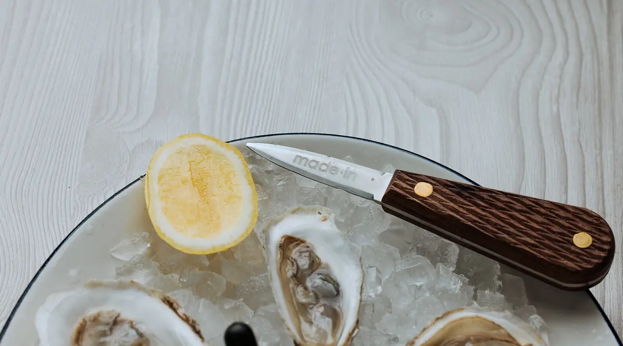 A close-up view of fresh oysters on ice with a slice of lemon and an oyster knife on a glass dish on a light wooden table.