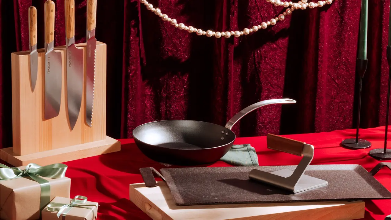 A set of kitchen knives on a stand, a frying pan on a cloth, and a sharpening stone on a cutting board, all displayed elegantly on a red tablecloth against a red velvet curtain backdrop.