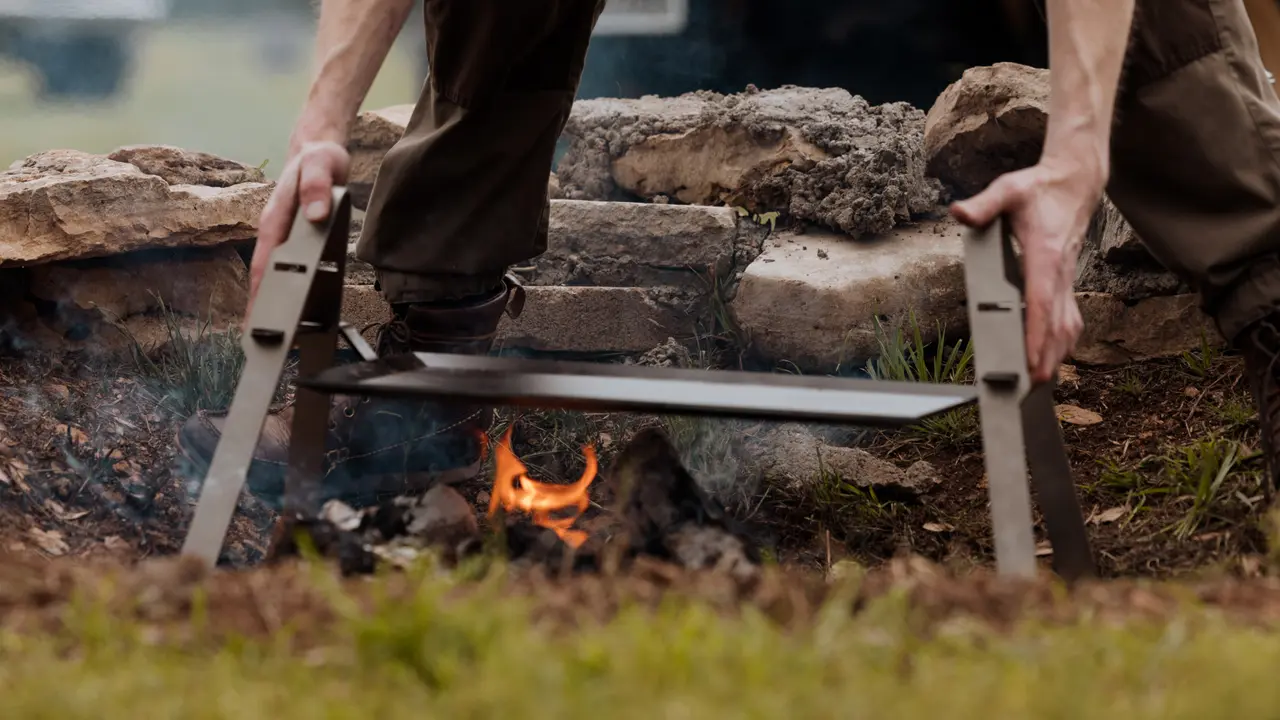 A person stands holding a long saw blade, preparing to cut a piece of wood over a small, open flame.