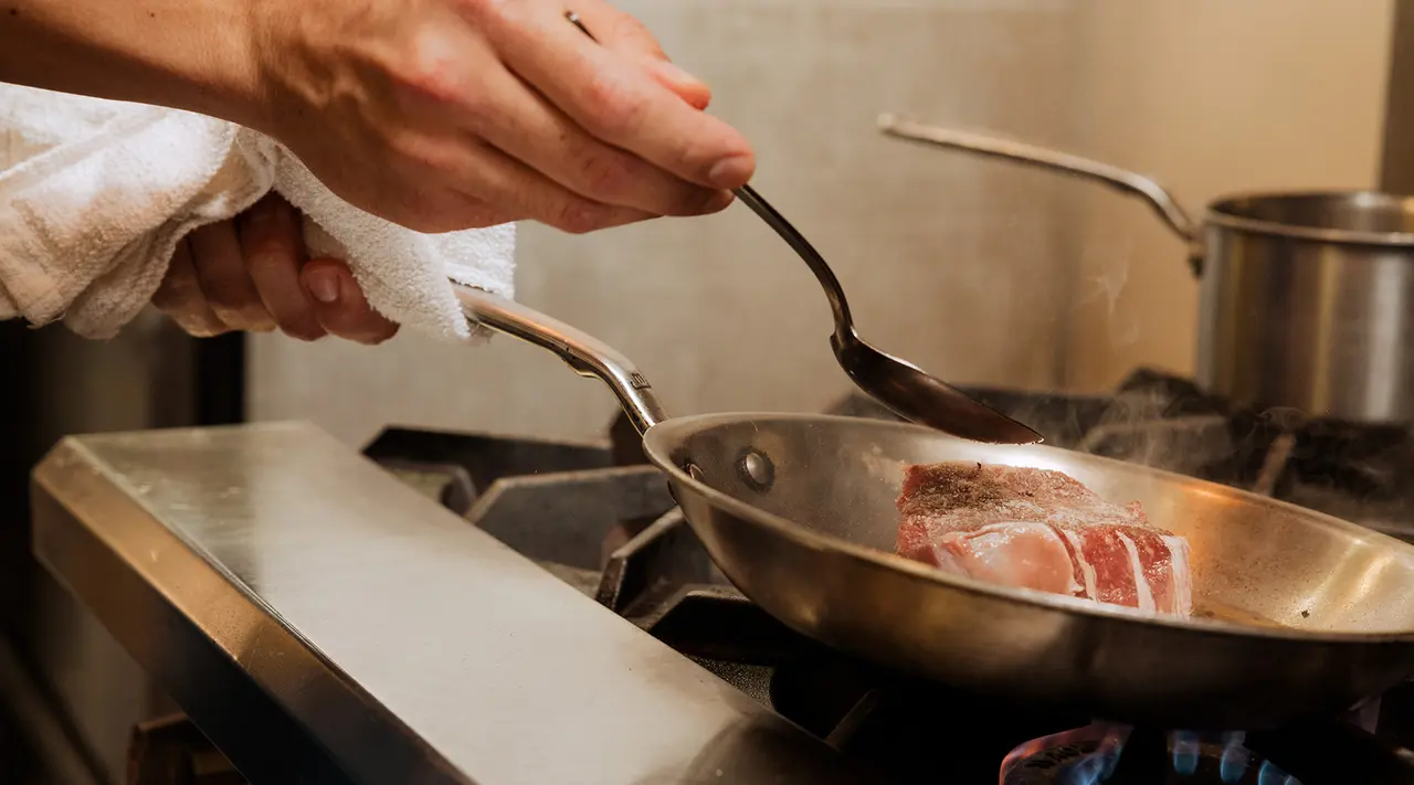 A person's hands using tongs to cook a piece of meat in a skillet over a gas stove.