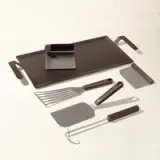 A set of griddle cooking tools, including two spatulas, a chopper/scrapper, and tongs, neatly arrayed beside a flat-top griddle with a drip tray.