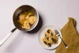 A serving of ravioli in broth with fresh basil leaves is presented in a white bowl beside a pot containing more ravioli.