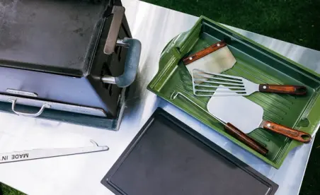 A portable grill is shown alongside a green tray containing grilling utensils and a black tray on a stainless steel surface.