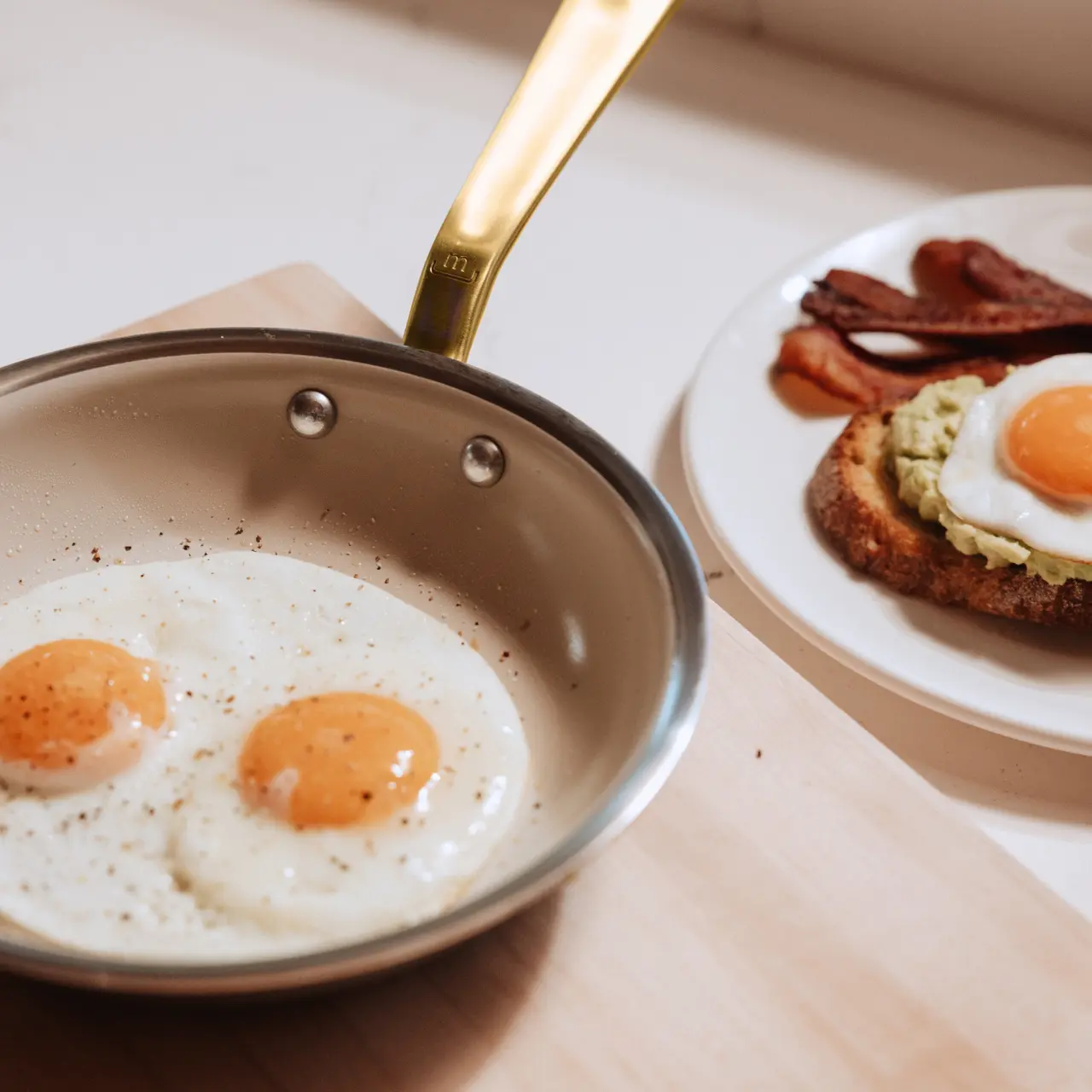 Two perfectly fried eggs with runny yolks sit in a small pan beside a plate of avocado toast topped with another egg and strips of bacon.